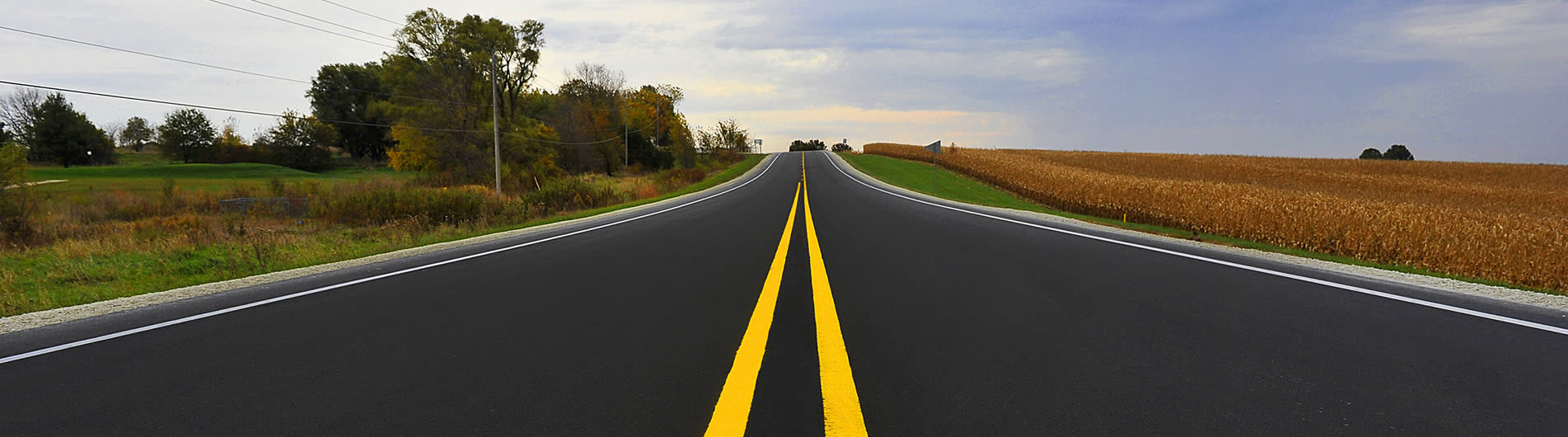 A newly paved two-lane Iowa highway with fresh double yellow centre lines, running between a cornfield and a stand of trees.